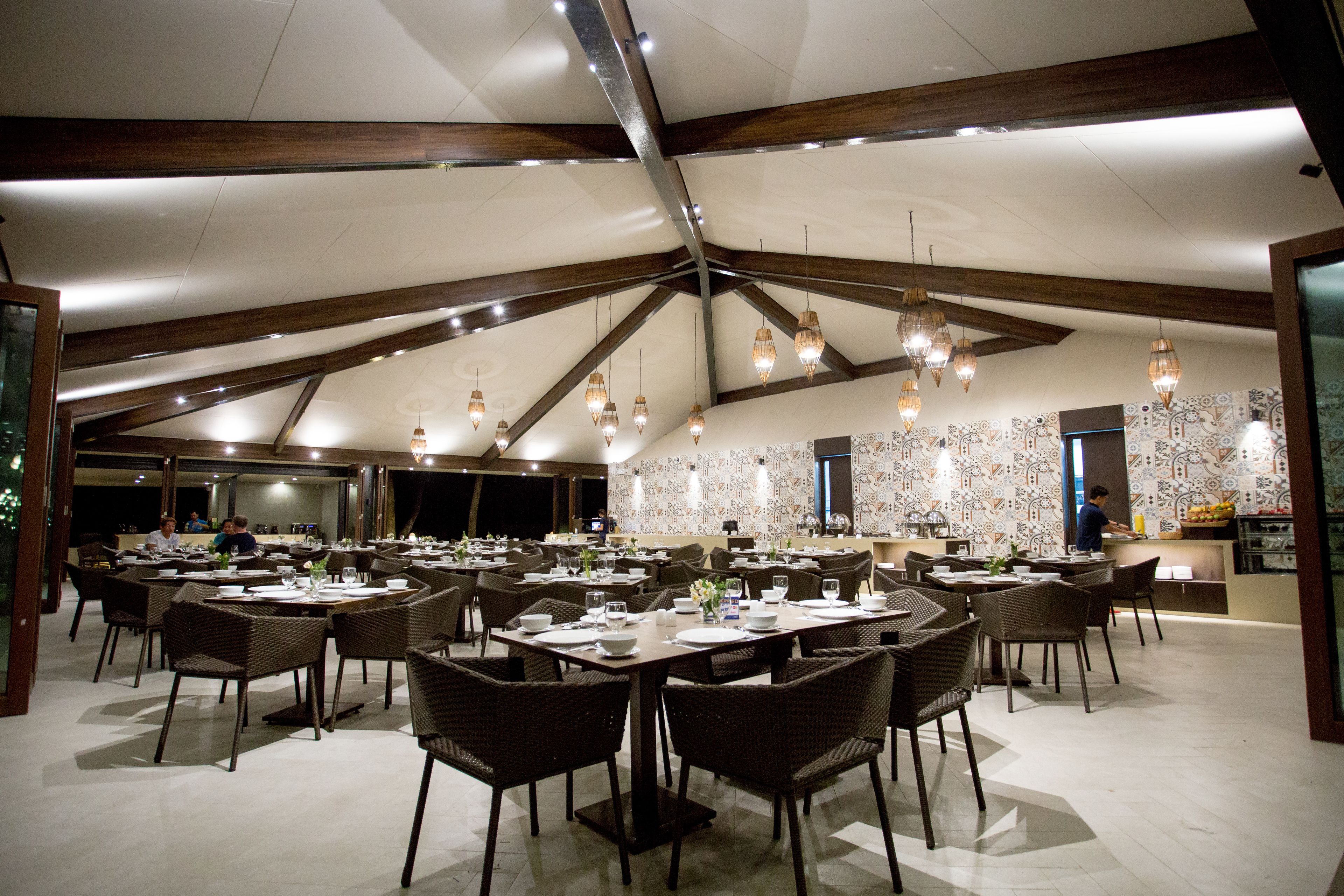 Un vaste espace dédié à la salle à manger, doté de hauts plafonds et de poutres apparentes, comprenant plusieurs tables et chaises, et un mur décoré d’un motif original en arrière-plan.