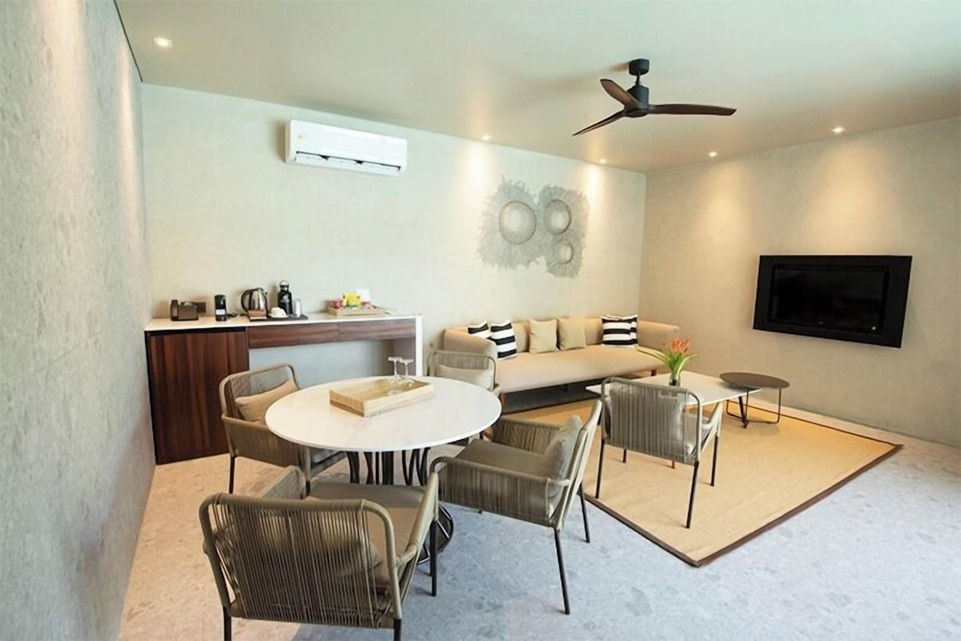 Una sala moderna con un sofá, una mesa de comedor y sillas. Hay un ventilador de techo, un televisor de pantalla plana montado en la pared y una cafetera sobre una encimera.