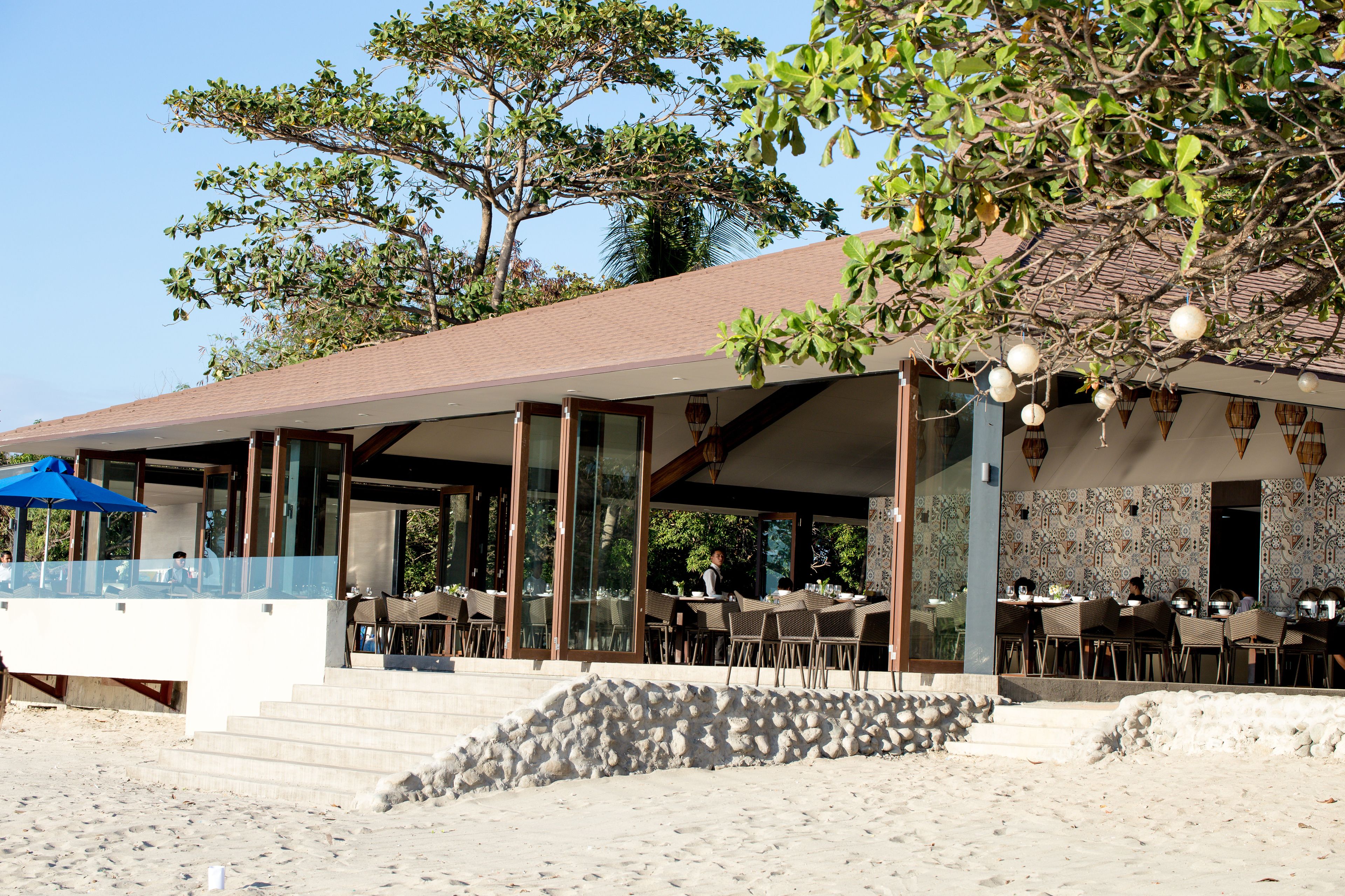 A beachfront restaurant with outdoor seating, a stone wall, and a covered patio area.