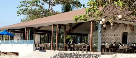 A beachfront restaurant with outdoor seating, a stone wall, and a covered patio area.