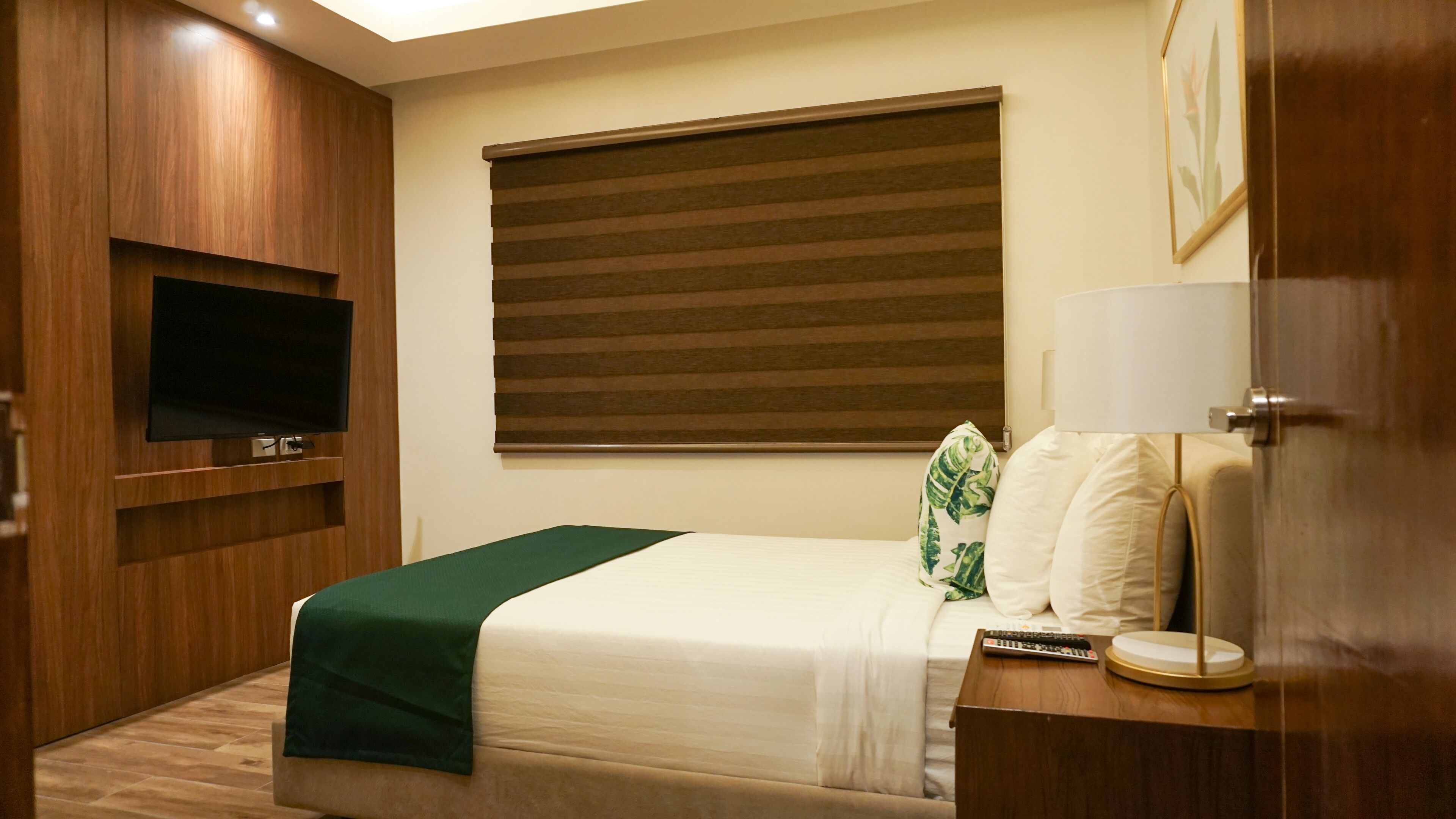 A hotel room with a bed, a TV mounted on the wall, a window with blinds, a bedside table with a lamp, and a door.