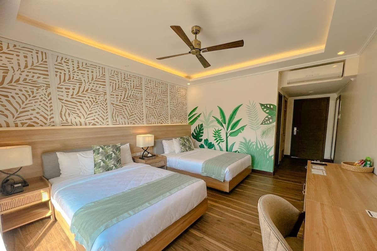 A hotel room with two beds, a ceiling fan, a wooden desk, and a wall with a leaf pattern.