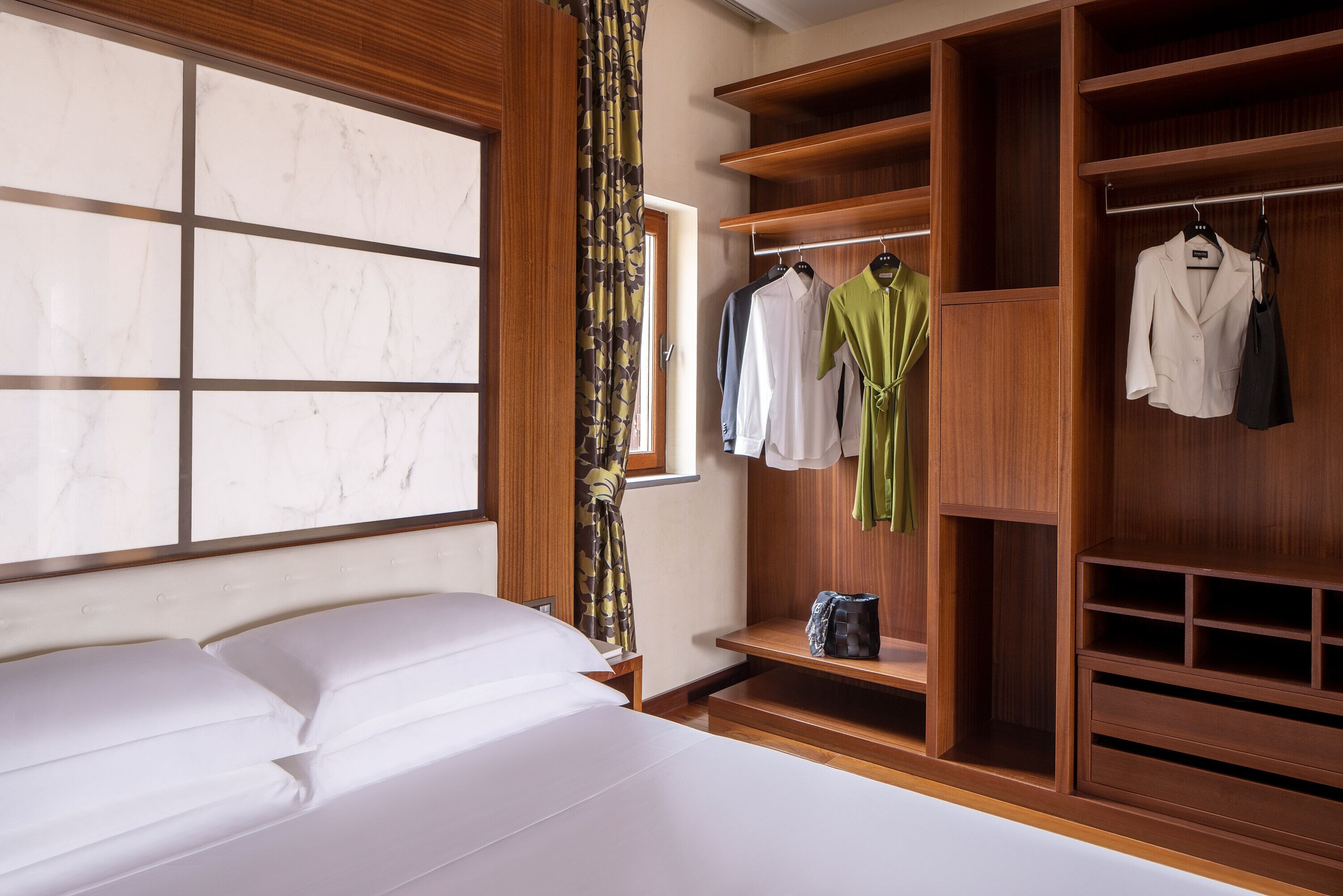 Une chambre d’hôtel avec un grand lit, une armoire en bois où sont suspendus des vêtements, et une fenêtre donnant sur un beau paysage.