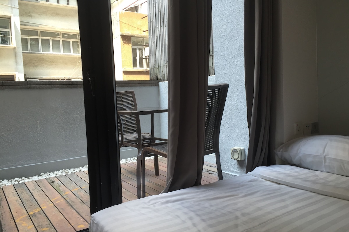 A hotel room with a bed, a small table with a chair, and a view of the outside balcony.
