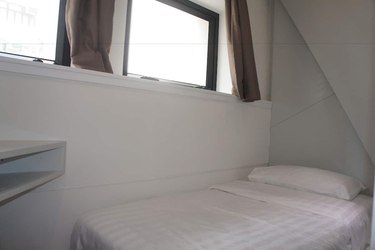 A small, single-bed room with a window and a white shelf.