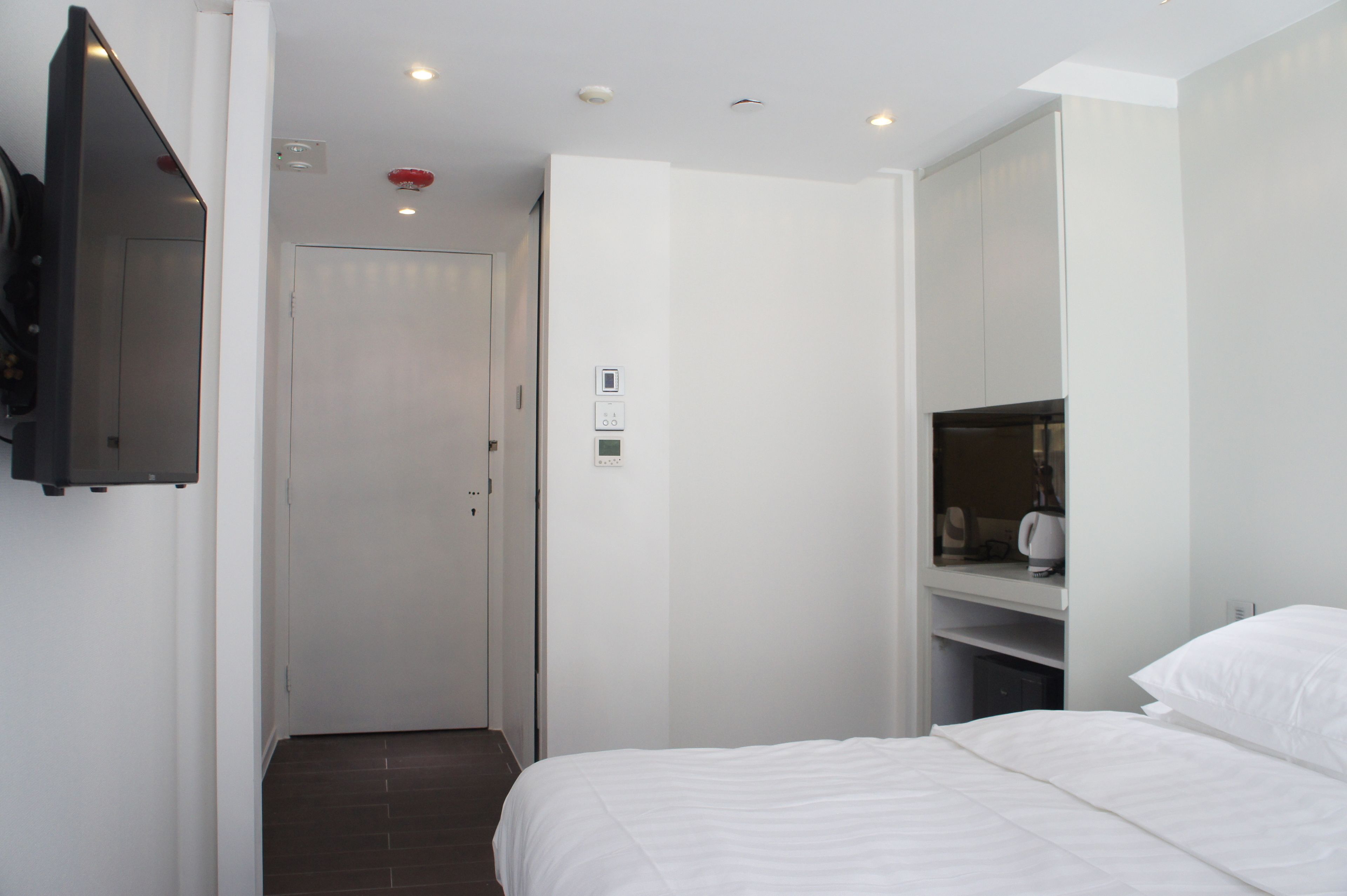 A hotel room with a bed, a TV mounted on the wall, a wardrobe, and a small kitchenette area.