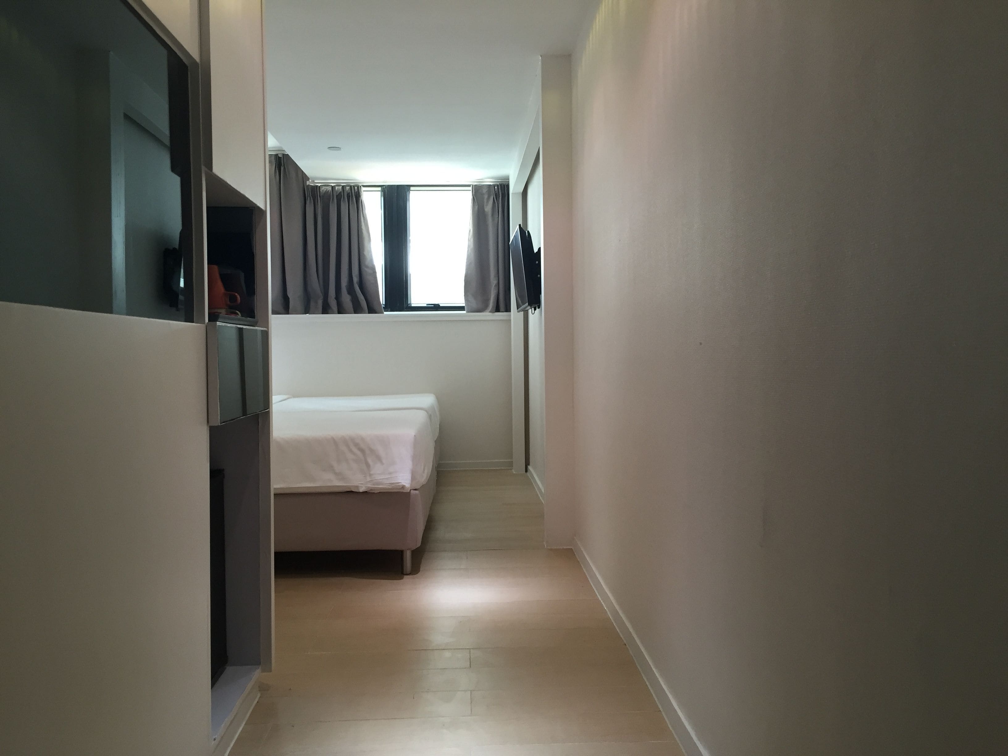 A narrow hallway with a bed at the end, a window with curtains, and a mirror on the left wall.