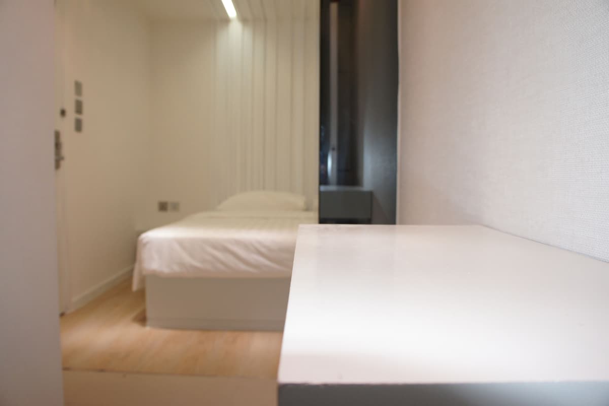 A small, narrow hotel room with a single bed and a small table.