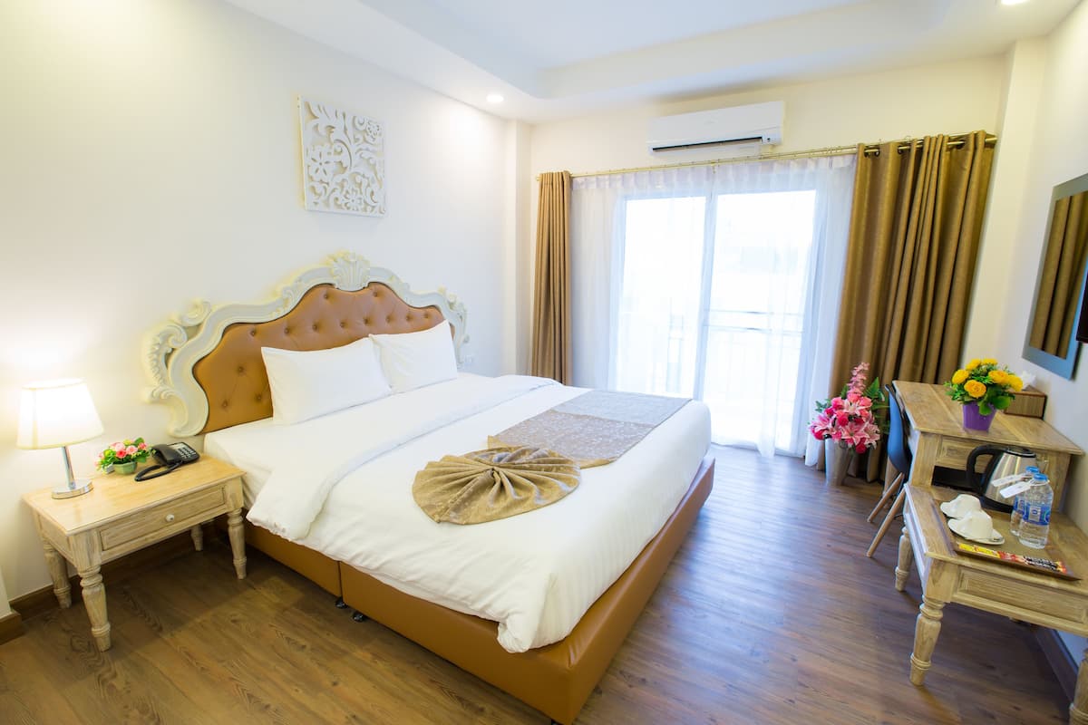 A neatly made bed with a headboard, a bedside table with a lamp and a phone, and a wooden desk with a mirror and a vase of flowers.