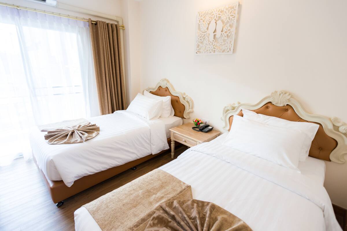 A hotel room with two beds, a nightstand with a phone and a small table, and a window with curtains.