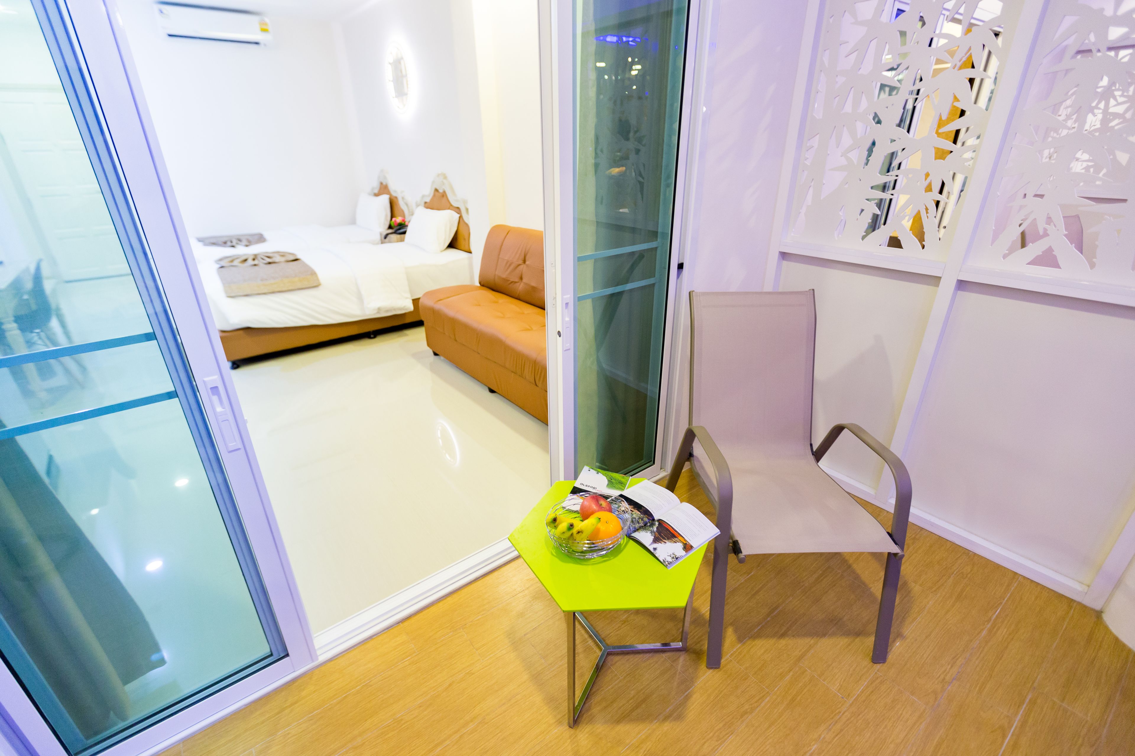 A modern hotel room with a glass door, a wooden floor, a green table with a fruit bowl, a magazine, and a chair.