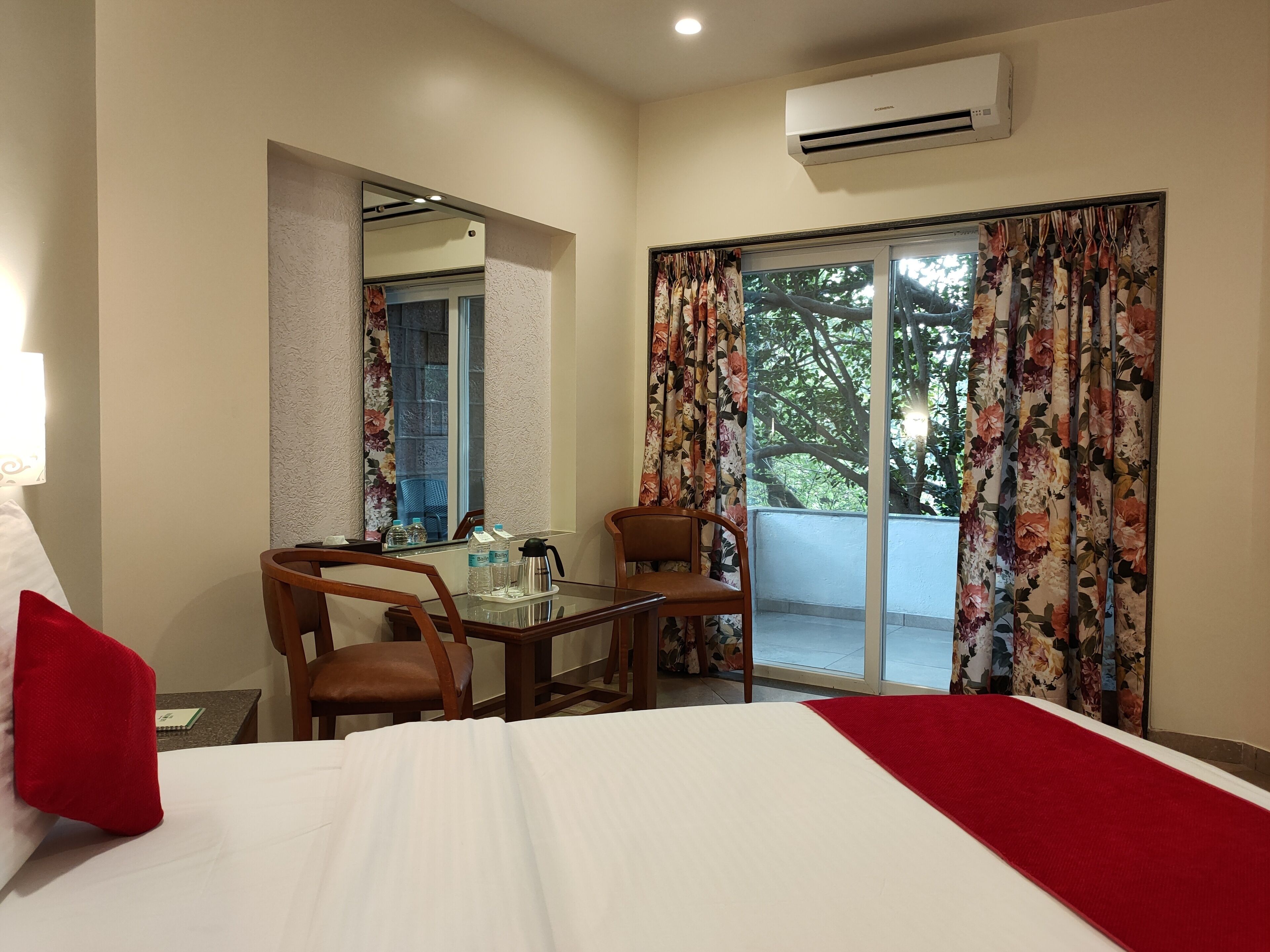 A hotel room with a bed, a table with chairs, a window with floral curtains, and an air conditioning unit.