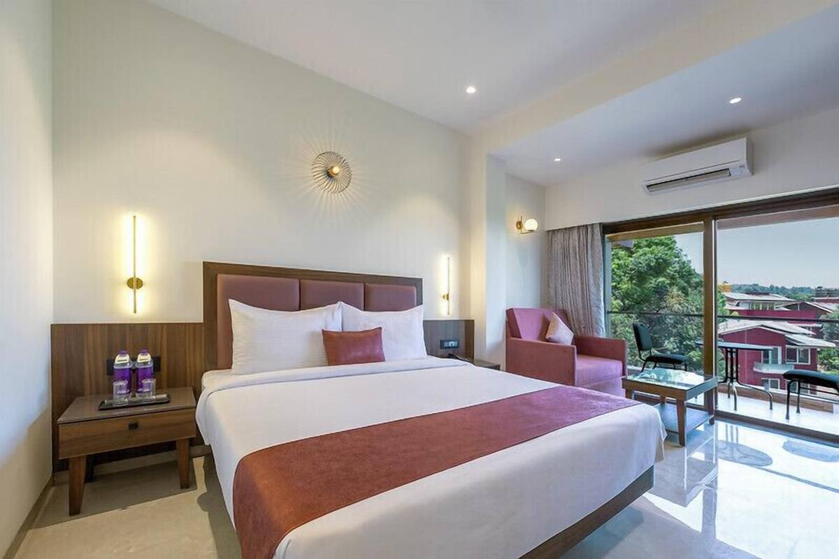 A hotel room with a large bed, a nightstand with a lamp, a chair, and a balcony with a view.