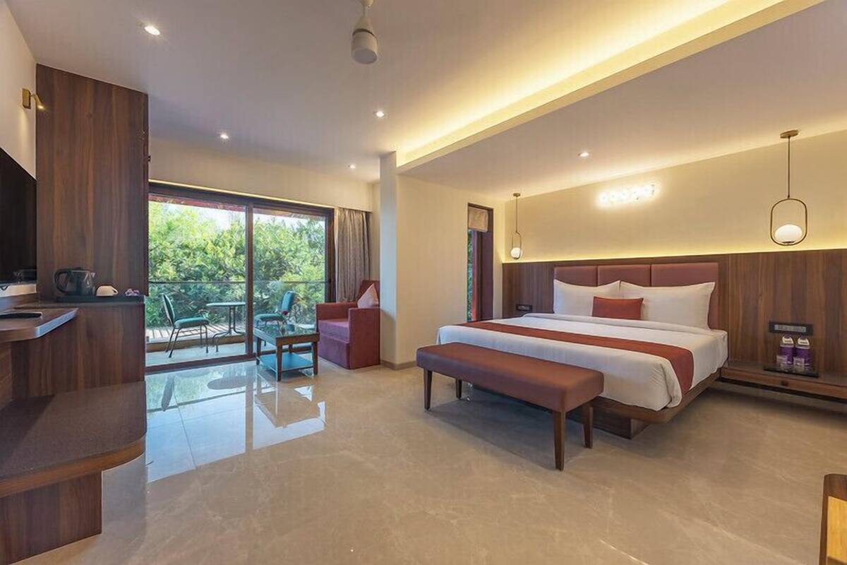 A modern hotel room with a large bed, a desk, and a balcony with a view of greenery.