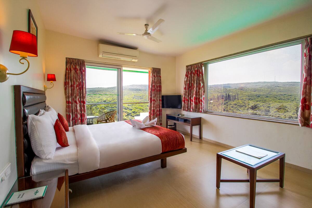 A hotel room with a bed, desk, TV, and large windows offering a view of the landscape.