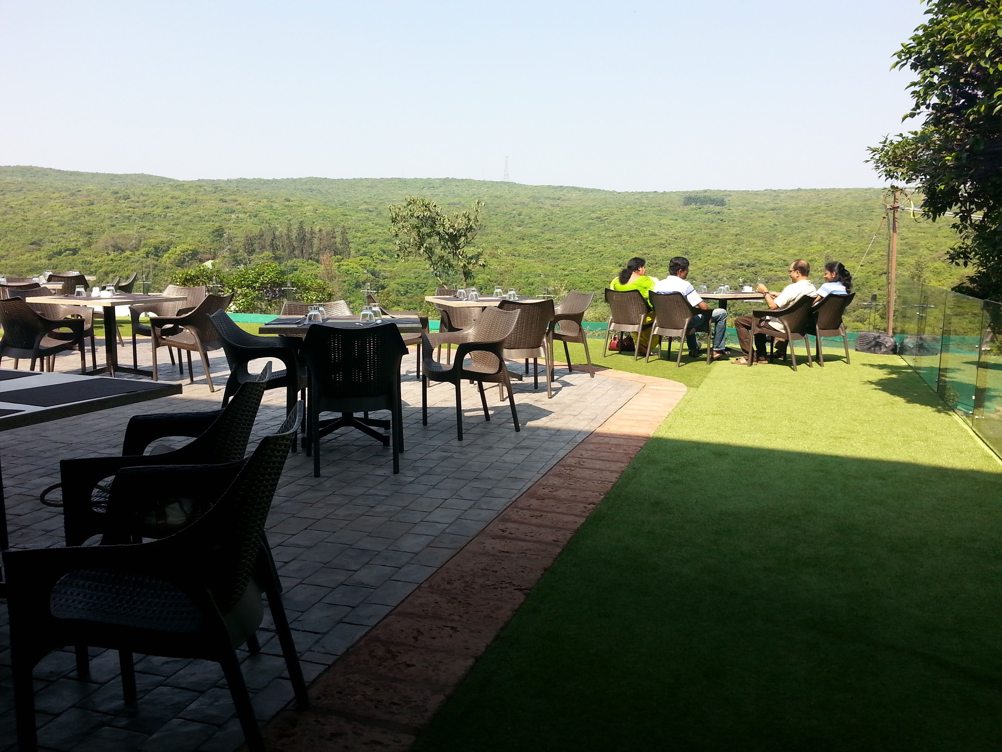 An outdoor dining area with tables and chairs, surrounded by a green lawn and hills in the background.