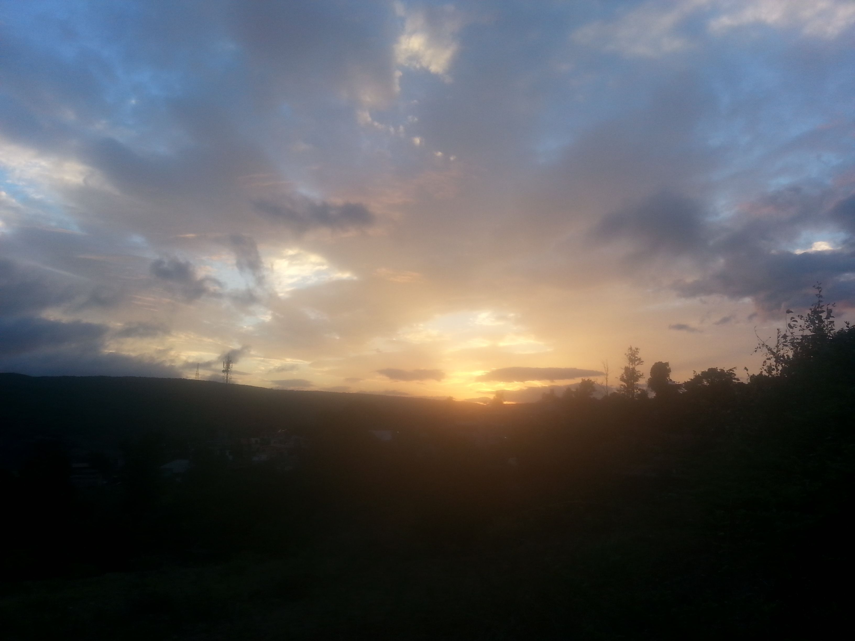 Een zonsondergang boven een heuvelachtig landschap, met silhouetten van bomen en een elektriciteitspaal.