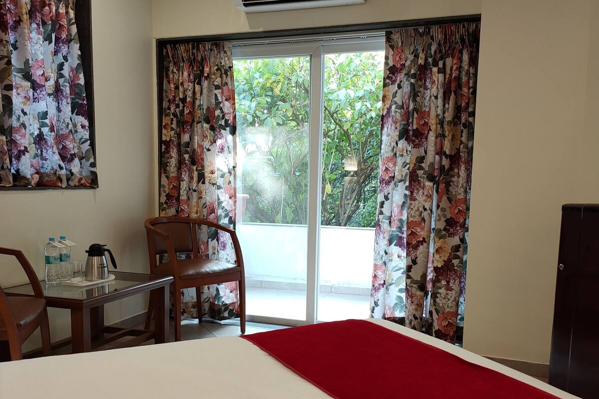 A hotel room with a bed, a table with a coffee maker, a chair, and a view of greenery through the window.