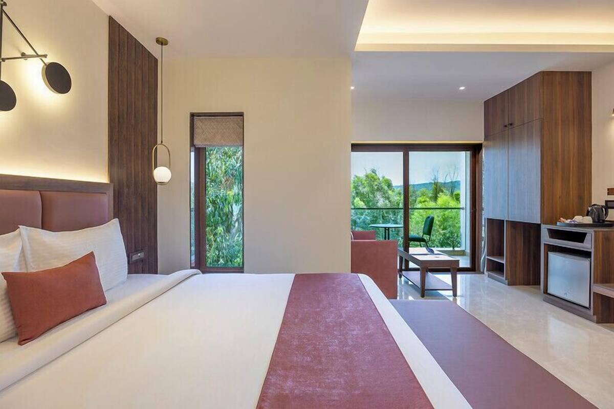 A modern hotel room with a large bed, a dining area, and a view of greenery.