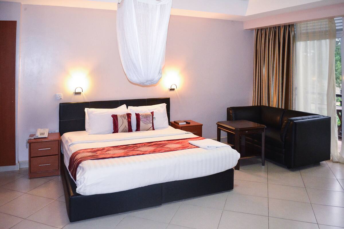 Quarto de hotel com cama grande, mesas de cabeceira, uma cadeira e janela com cortinas.