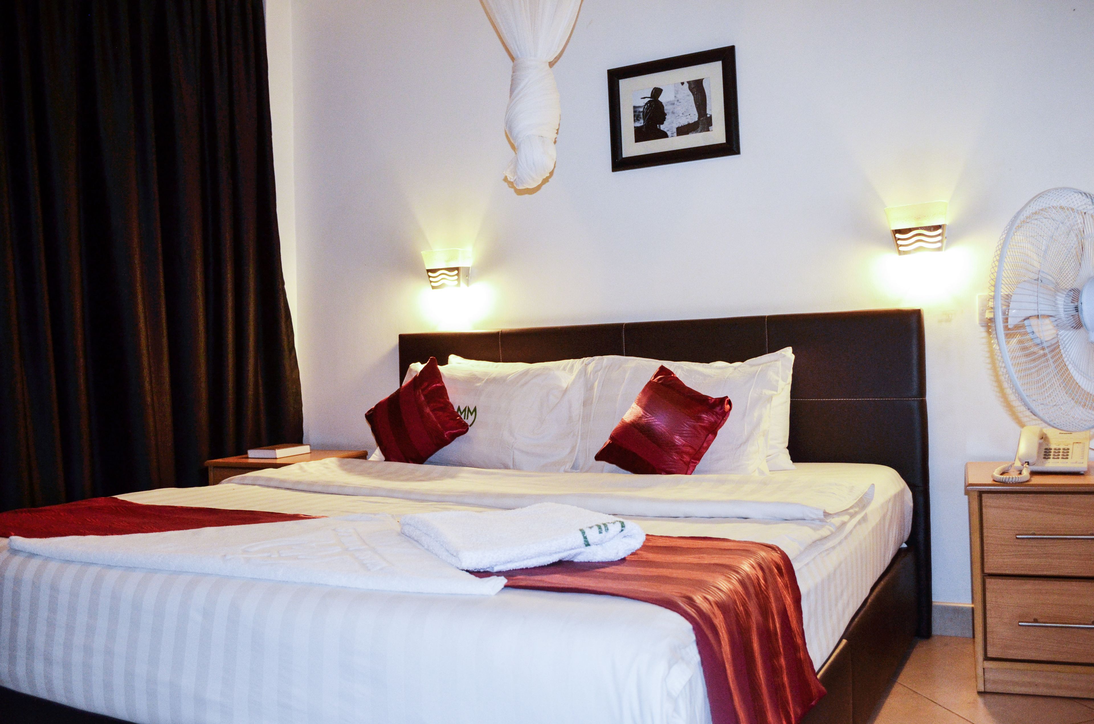 A neatly made bed with white and red bedding, two bedside lamps, a wall-mounted picture, and a bedside fan.