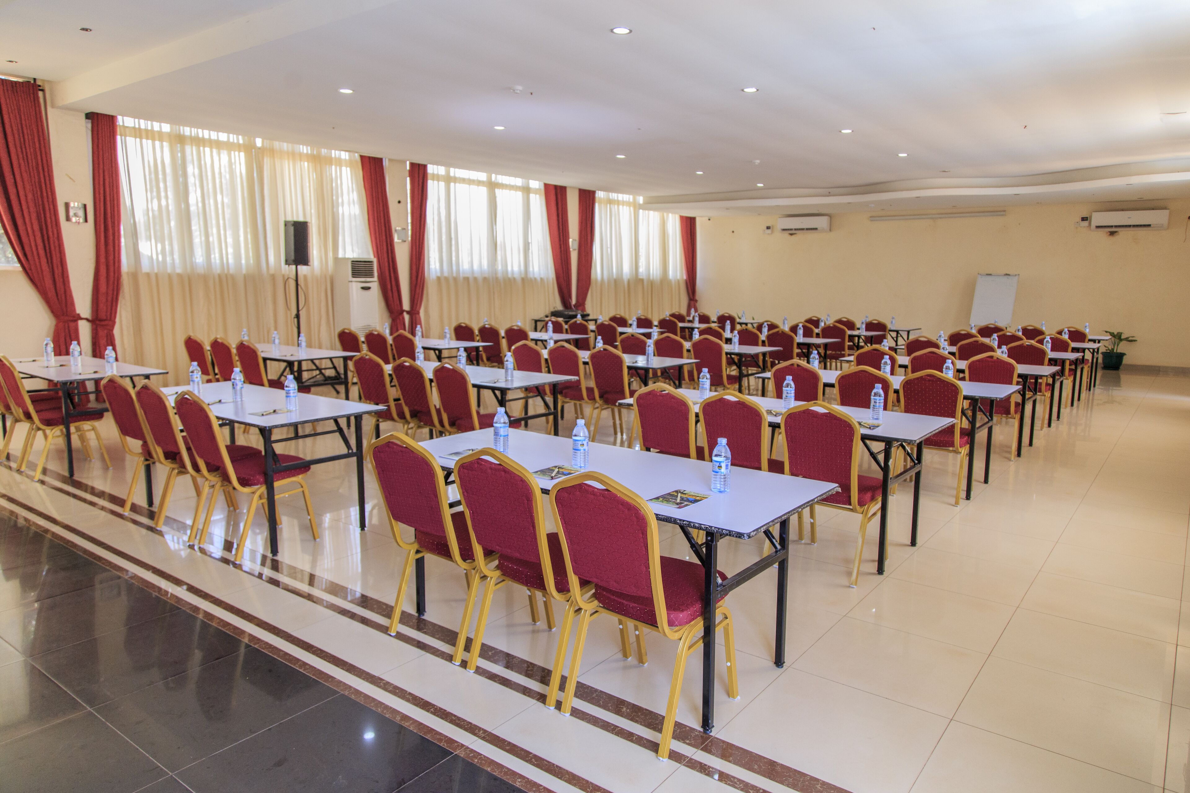Une salle de conférence avec des rangées de tables et de chaises, équipée de bouteilles d’eau et d’un tableau blanc.