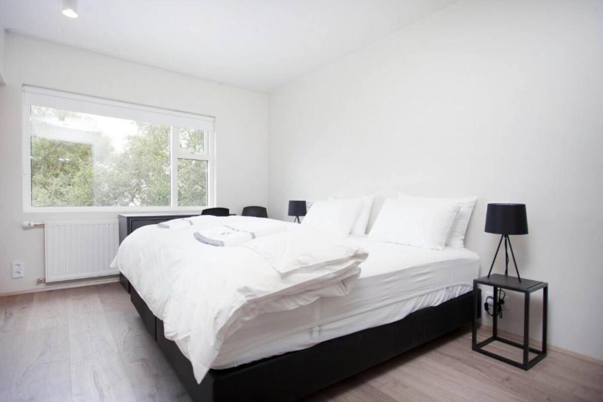 A bedroom with a large bed, two bedside tables with lamps, a window with a view of trees, and a radiator under the window.