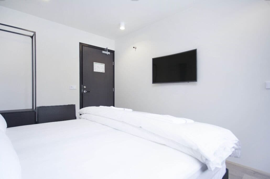 A hotel room with a bed, a door, a flat-screen TV mounted on the wall, and a light fixture.