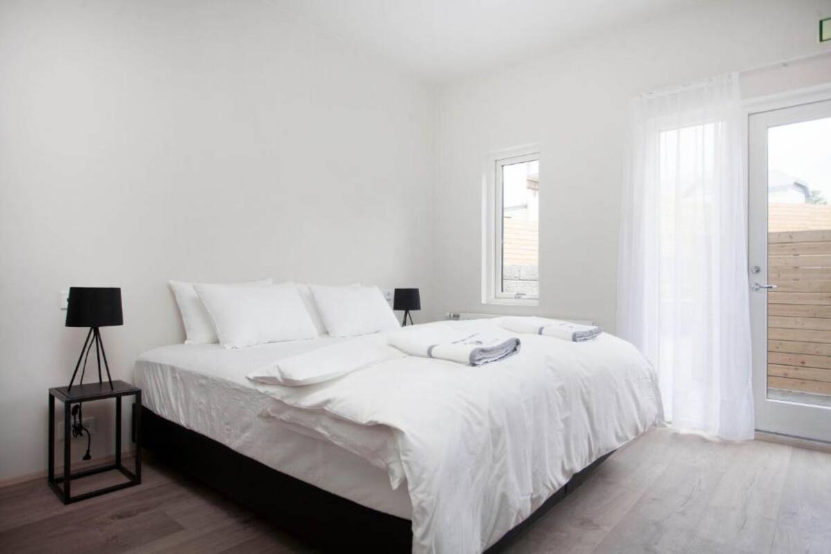 A bedroom with a bed, two bedside lamps, a window, and a view of a wooden deck.