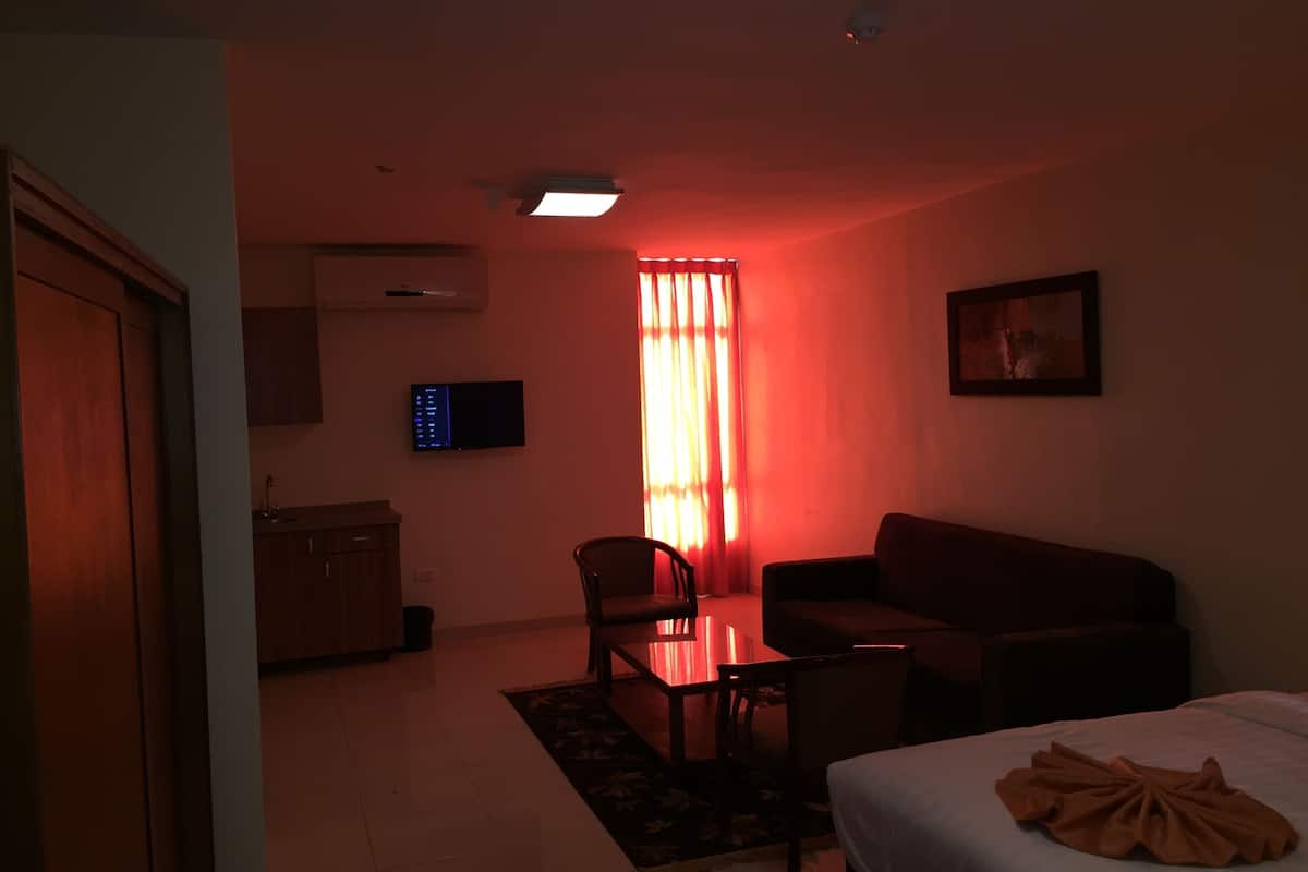 A dimly lit room with a red hue, a flat-screen TV mounted on the wall, a sofa, a dining table with chairs, and a bed with a blanket.