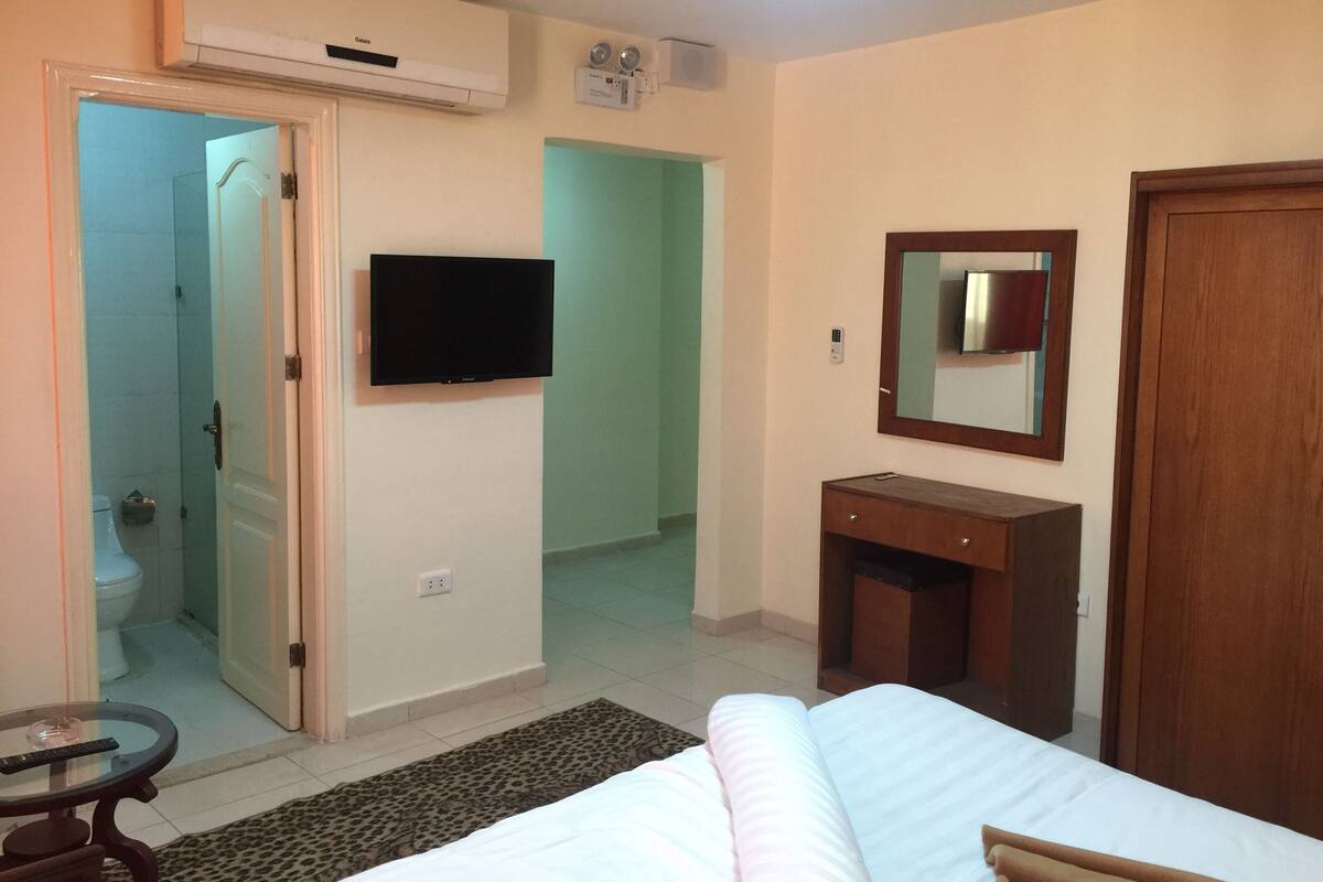 A hotel room with a bed, a bedside table, a TV mounted on the wall, a mirror, and an air conditioning unit.
