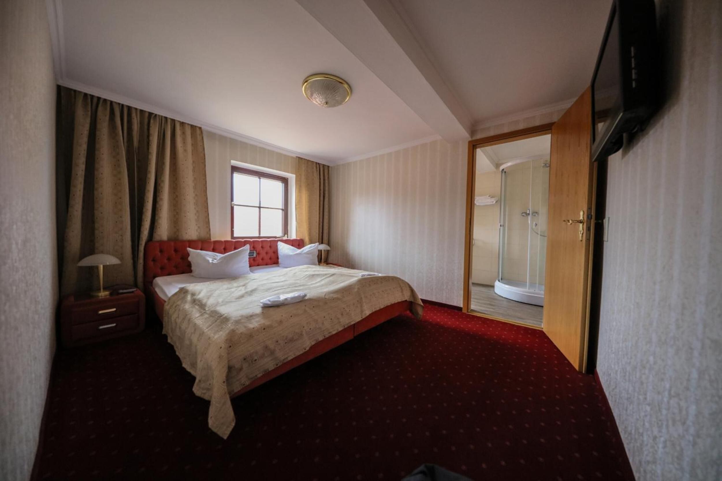 A hotel room with a large bed, a bedside table with a lamp, a window with curtains, a shower area, and a television mounted on the wall.