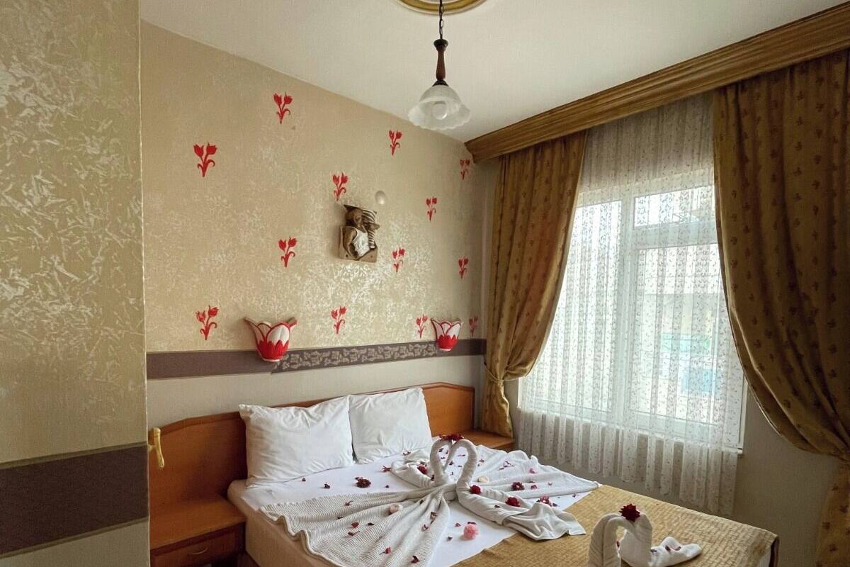 A hotel room with a bed, a nightstand, a wall-mounted shelf with decorative items, a ceiling light, and a window with curtains.