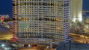 Un hotel de varios pisos con numerosas ventanas, un estacionamiento y un edificio cercano con techos iluminados.