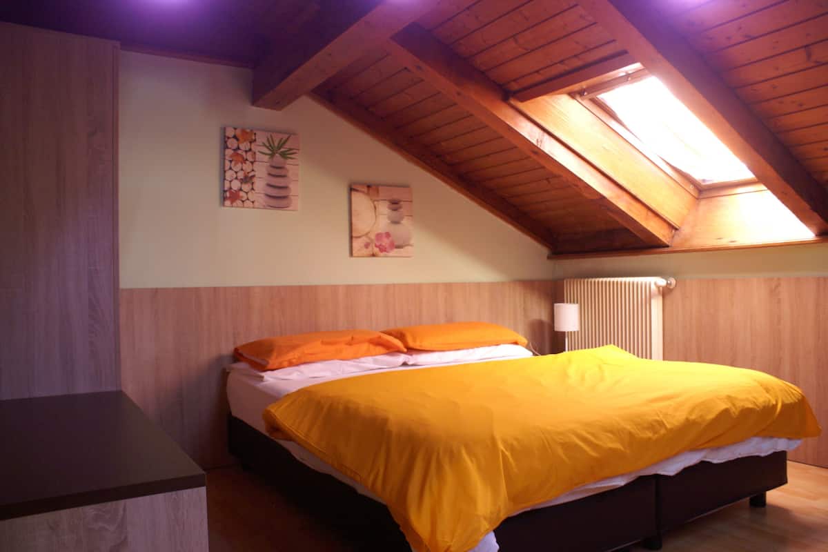 A bedroom with a sloped ceiling, wooden beams, a bed with orange bedding, and two wall-mounted artworks.