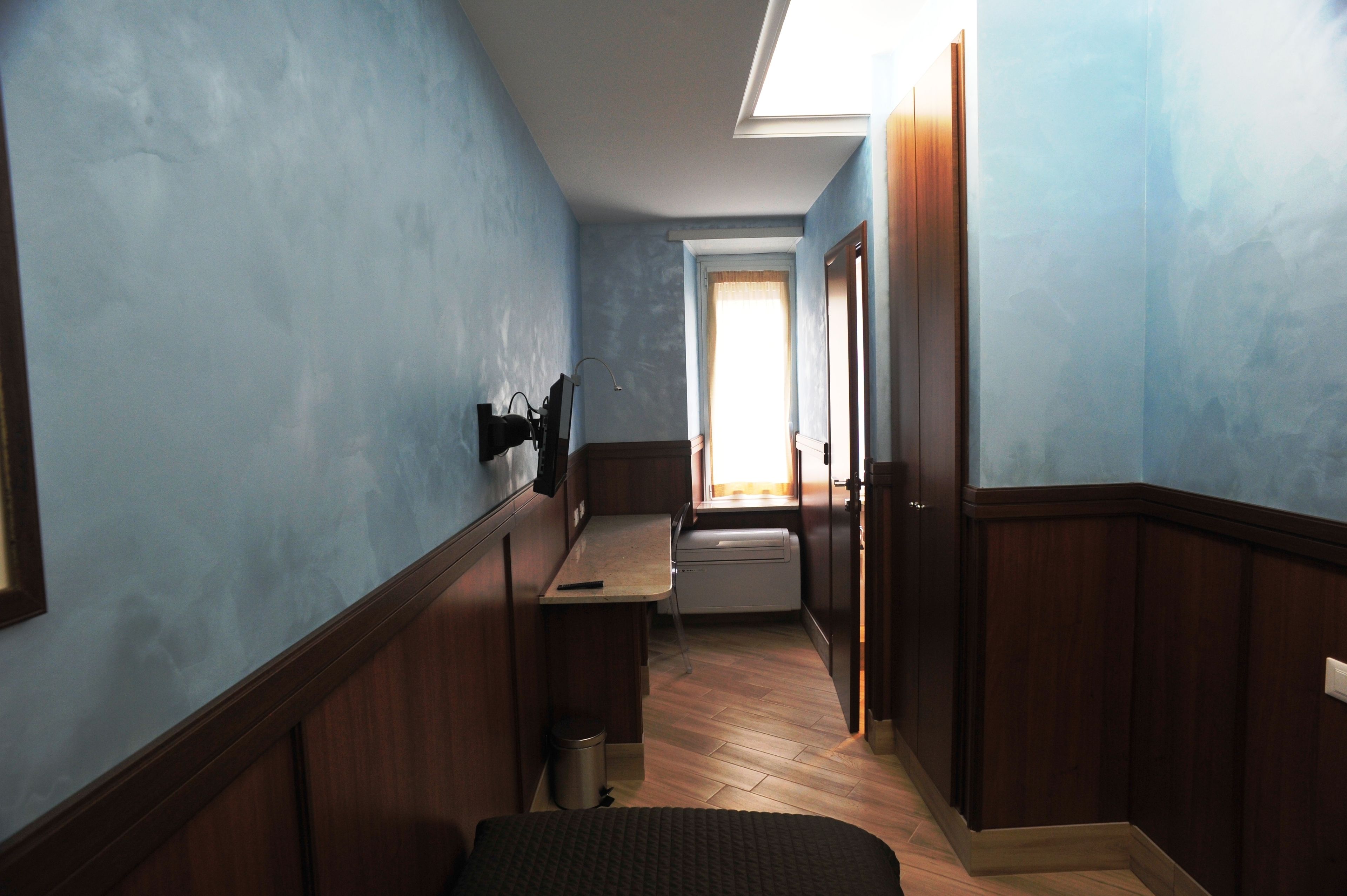 A narrow hallway with wooden paneling, a small desk with a chair, a trash can, and a window with curtains.