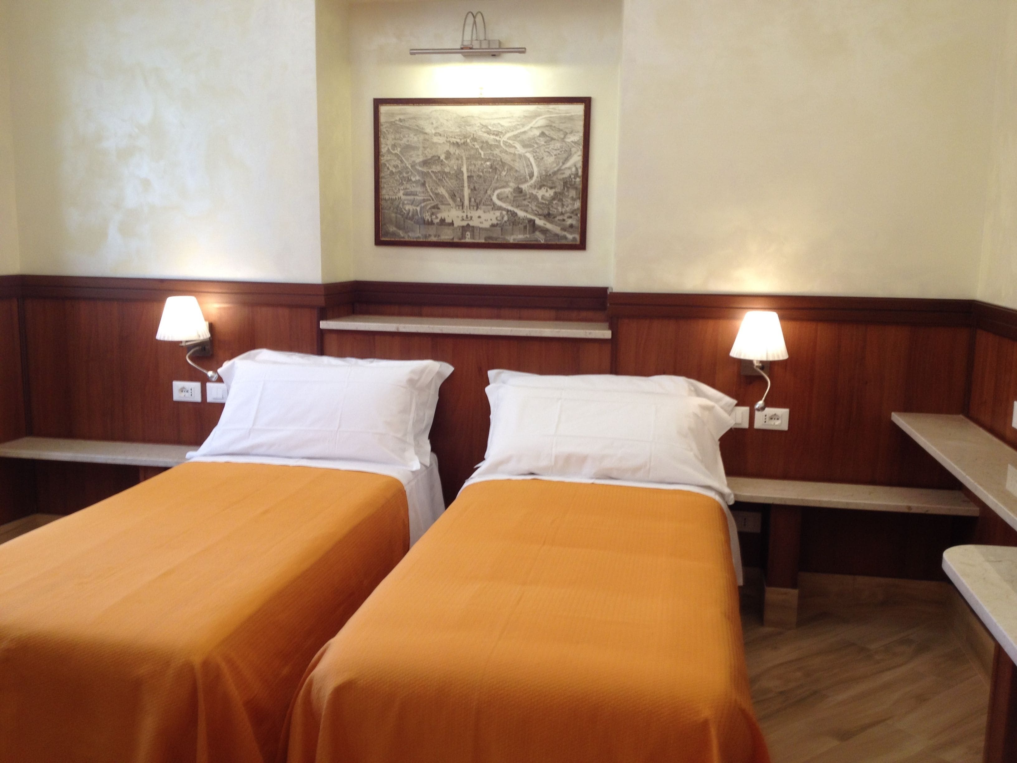A hotel room with two single beds, each with a wooden headboard and a white pillow. There is a framed picture on the wall above the beds, and two bedside lamps on either side.