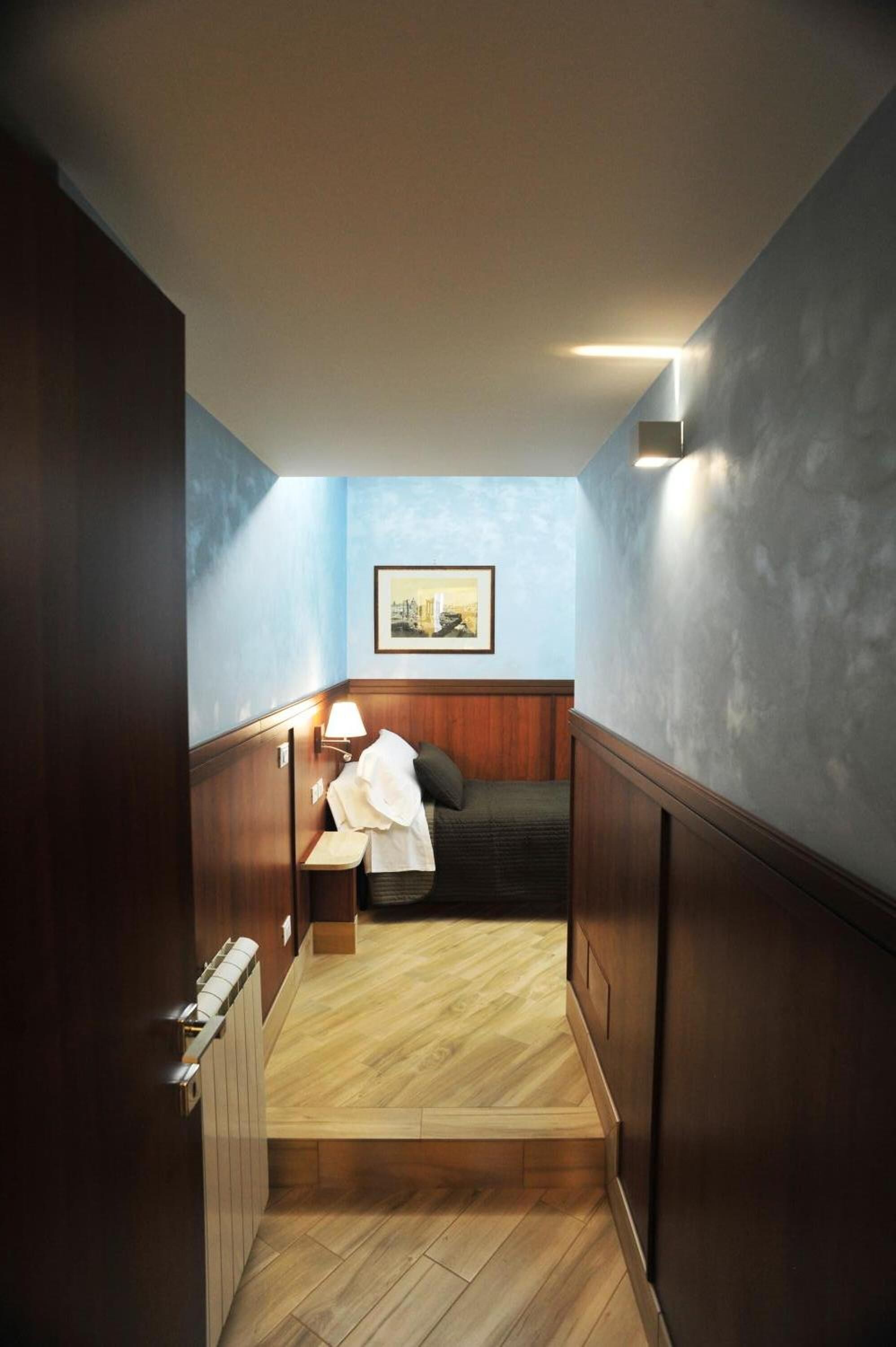 A narrow hallway with wooden flooring leading to a bedroom with a bed, a nightstand, and a wall-mounted lamp.
