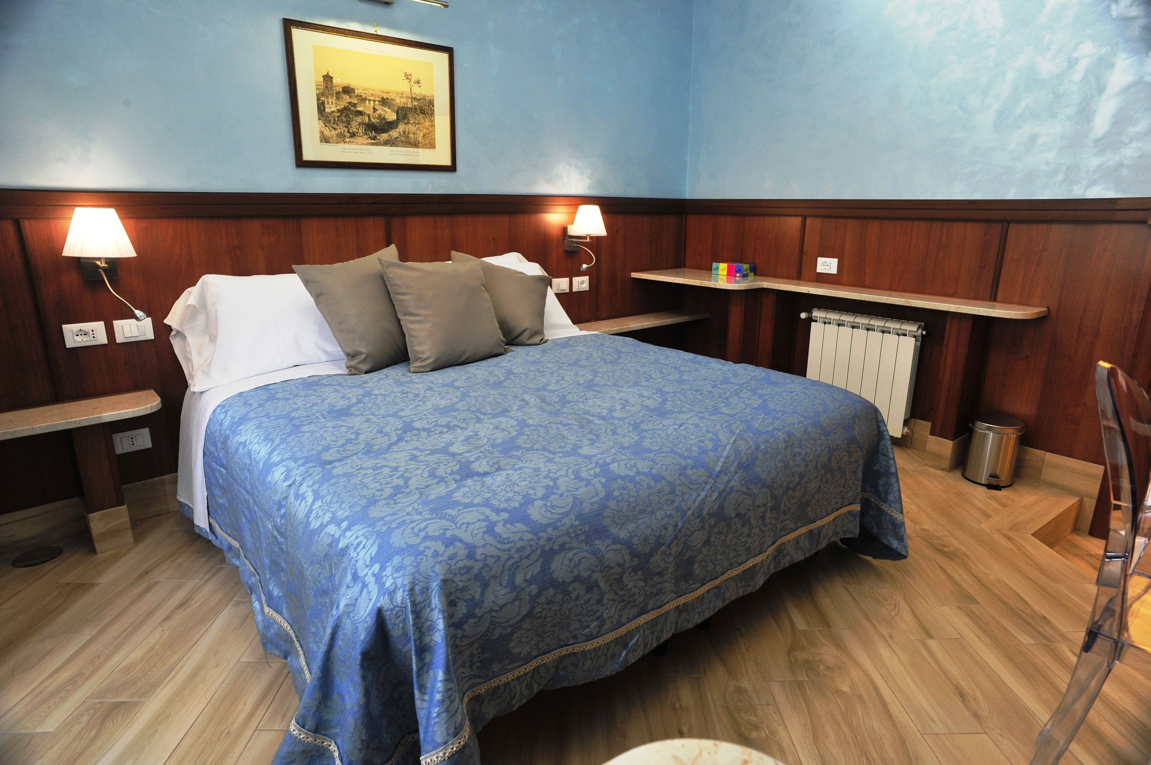 A hotel room with a large bed, two bedside lamps, a framed picture on the wall, and a small shelf with items.