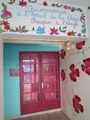 Un espace d’accueil avec un mur de couleur turquoise et une porte rouge, décoré de motifs floraux et agrémenté d’une pancarte de bienvenue.