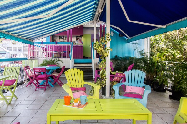 A colorful outdoor seating area with vibrant chairs and tables, surrounded by potted plants and a blue awning.
