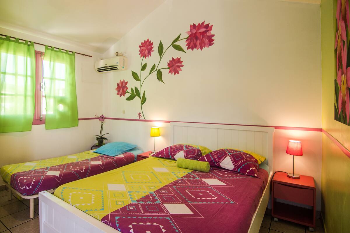 A room with two beds, a wall-mounted air conditioner, a window with green curtains, and a bedside lamp.