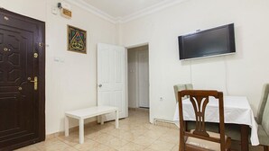 A room with a dark wooden door, a white bench, a television mounted on the wall, and a dining table with chairs. - Hotel Sufi (Istanbul)