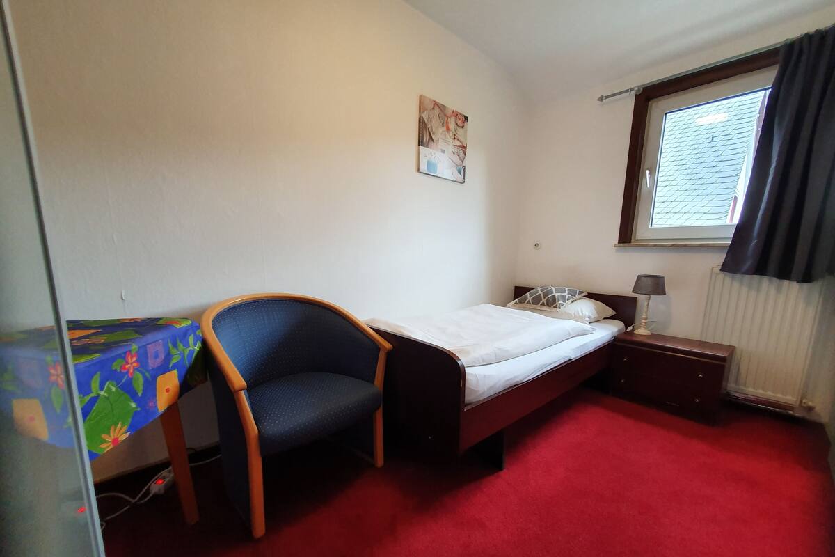 A small, neatly arranged room with a bed, a chair, a table, and a window.