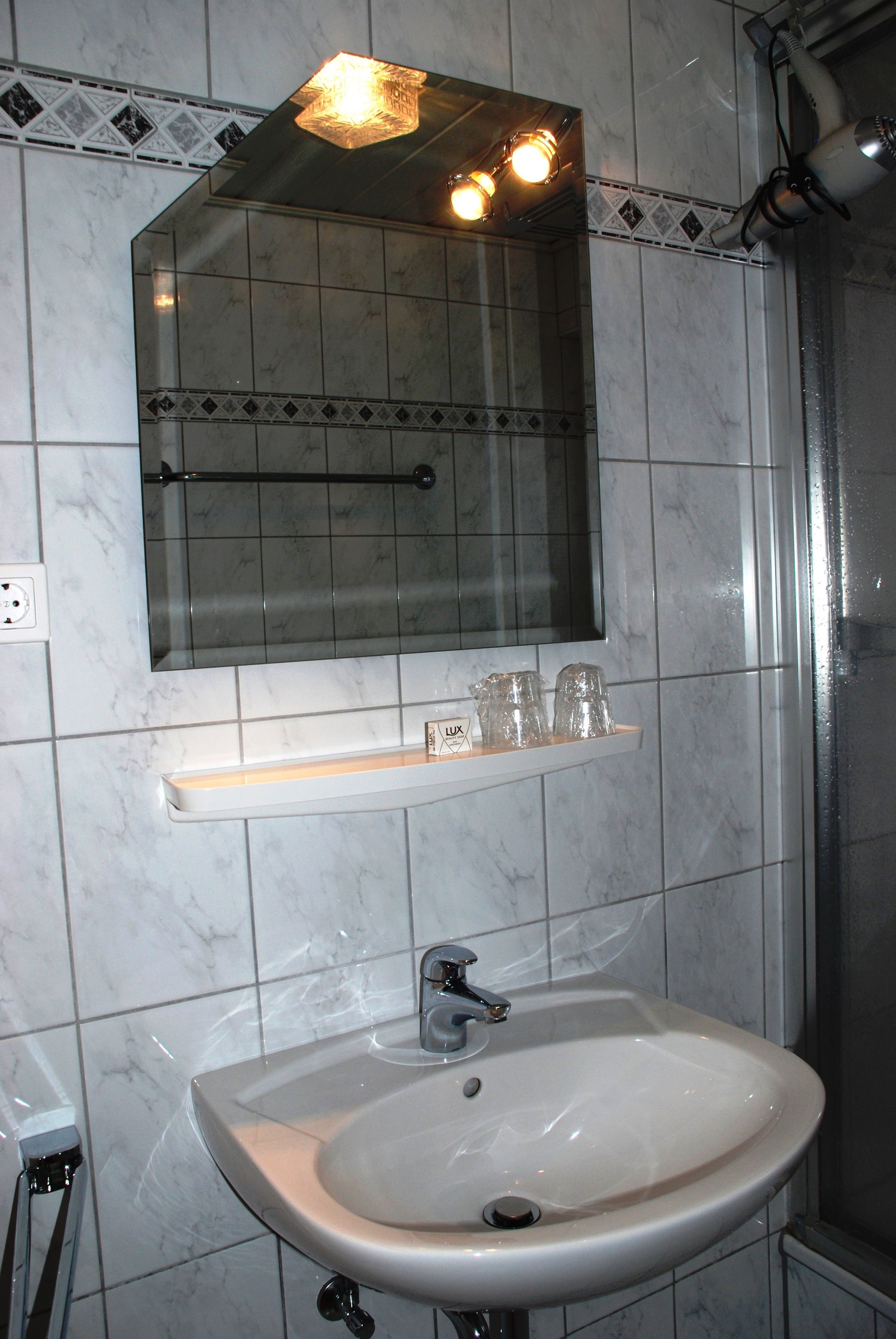 Un baño con un lavamanos blanco, un espejo encima y un estante con tres vasos de vidrio.