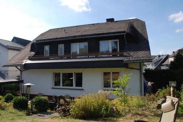 Une maison à deux étages, avec un toit sombre, des murs blancs et un balcon. On y trouve un jardin avec différentes plantes et une chaise à l’extérieur.