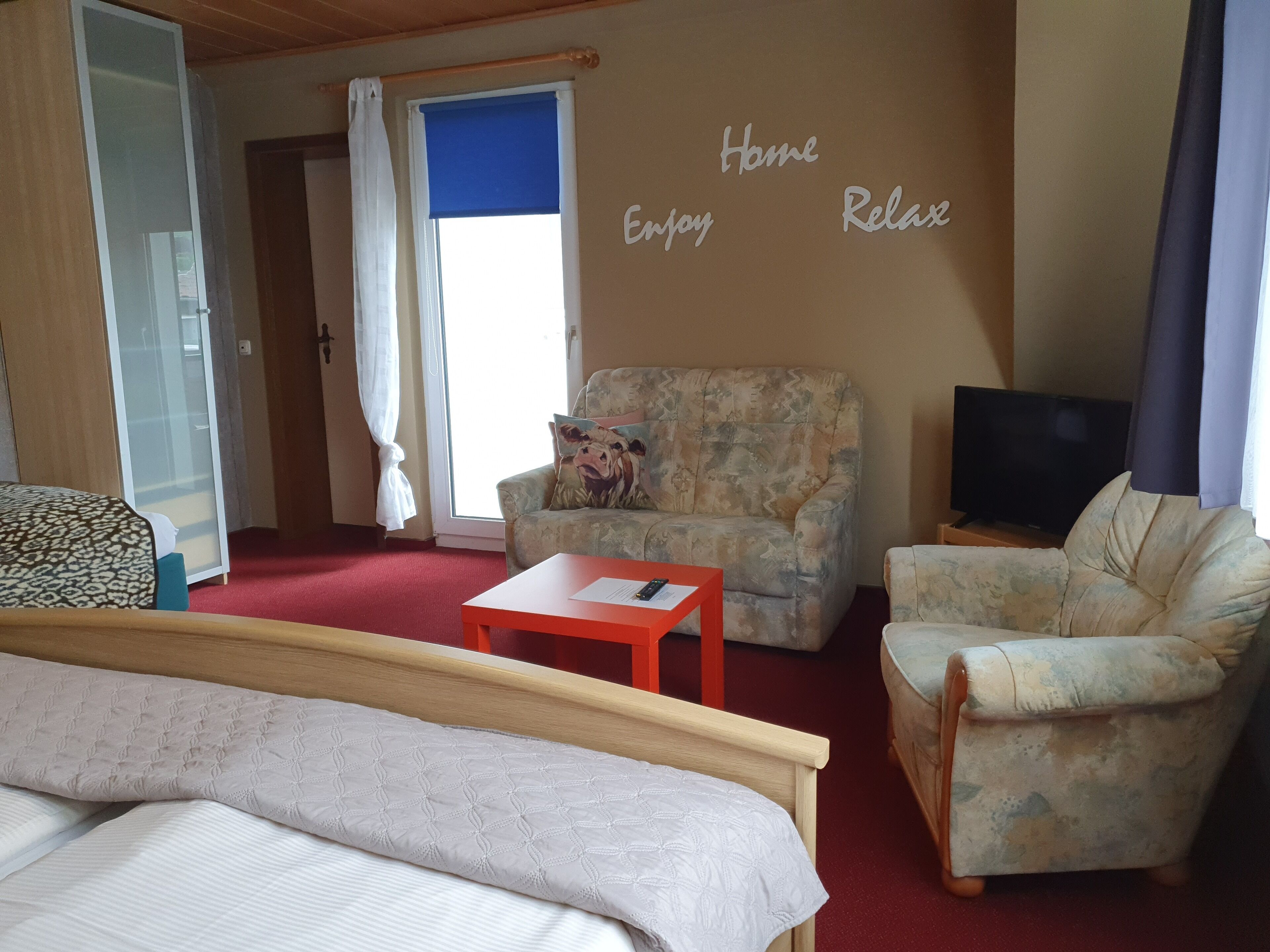 Una habitación con una cama, dos sillones, un sofá, una mesita, un televisor y una pared con la inscripción 'Home Enjoy Relax'.