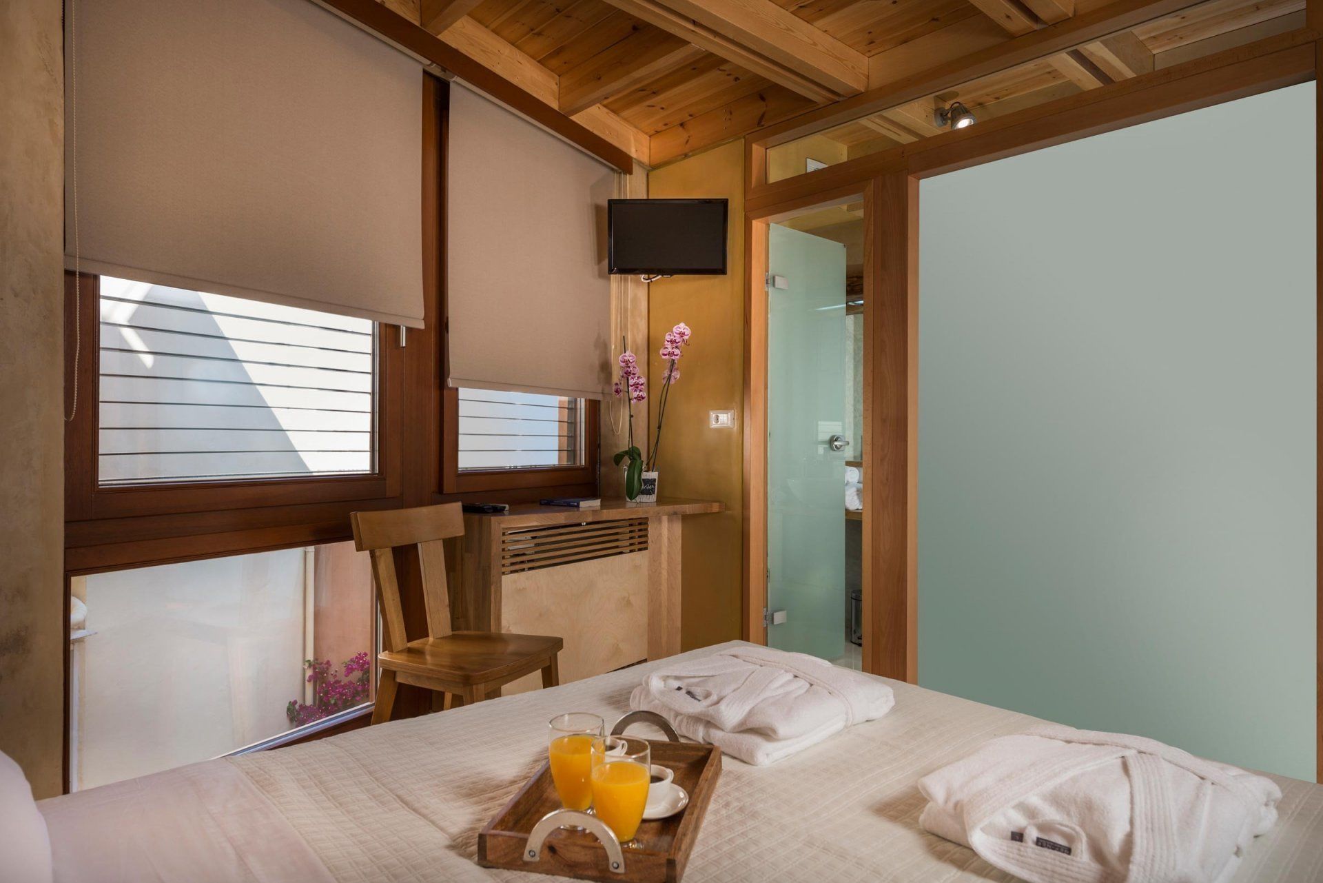 Une chambre d’hôtel avec un lit, une chaise en bois, un téléviseur fixé au mur et une fenêtre avec des stores.