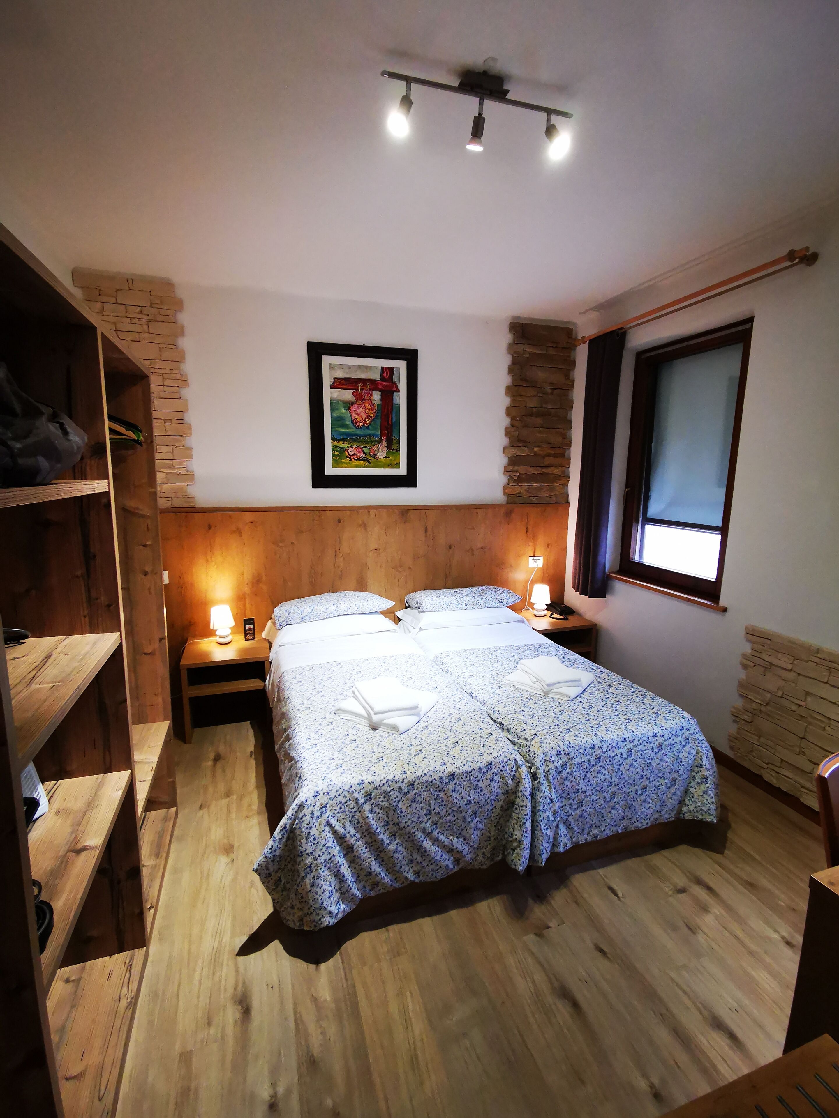 A bedroom with a wooden headboard, two beds with floral bedding, a nightstand with a lamp, a framed picture on the wall, and a window with a view.