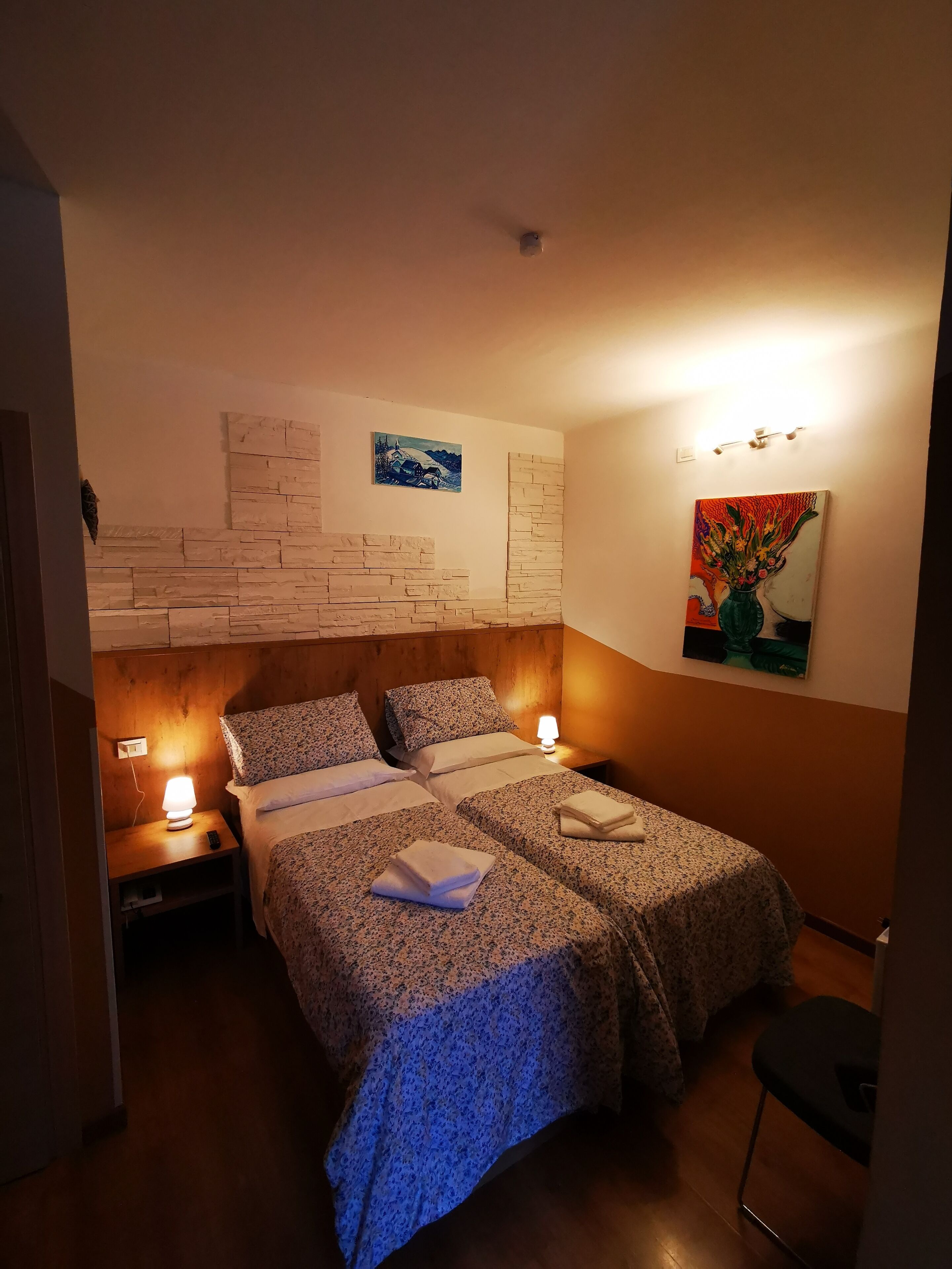 Una cama doble con ropa de cama floral, una mesita de noche con lámpara, un cuadro colgado en la pared y una pequeña mesa con un jarrón.